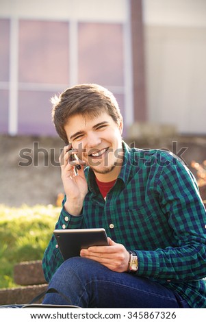 Smiling young man taping on tablet and speaking on mobile phone in a city on stair .Young smiling student  outdoors  with tablet and mobile phone.Life style.City