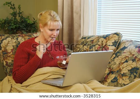 Woman concentrating while working at home on the couch. Cup of tea and glasses in hand with feet and legs covered with a blanket.