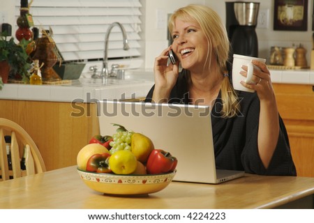 Woman smiling, in her kitchen on cell phone sitting in front of laptop.