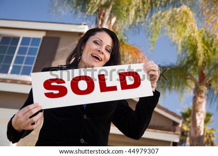 Happy Attractive Hispanic Woman Holding Sold Sign In Front of House.