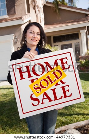 Happy Attractive Hispanic Woman Holding Sold Home For Sale Sign In Front of House.
