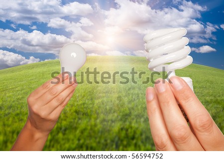 Female Hands Holding Energy Saving and Regular Light Bulbs Over Arched Horizon of Grass Field, Clouds and Sky.