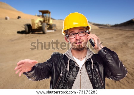 Young Construction Worker on Cell Phone in Dirt Field with Tractor in the Background.