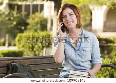 Smiling Young Pretty Female Student Outside on Cell Phone with Backpack and Books Sitting on Bench.