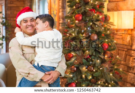 Hispanic Armed Forces Soldier Wearing Santa Hat Hugging Son In Front Christmas Tree.