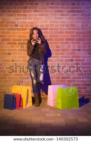 Pretty Mixed Race Young Adult Woman with Shopping Bags Using Her Cell Phone Against a Brick Wall - Plenty of Copy Space.