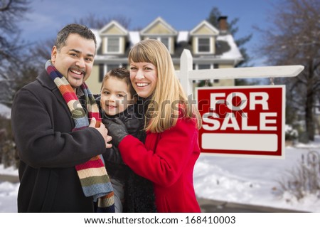 Warmly Dressed Young Mixed Race Family in Front of Home For Sale Real Estate Sign and House with Snow On The Ground.
