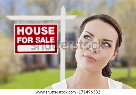 Thoughtful Pretty Mixed Race Woman In Front of Home and House For Sale Real Estate Sign Looking Up and to the Side.