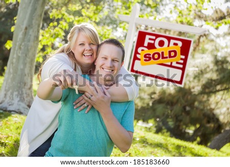 Happy Couple Holding House Keys In Front of Sold Real Estate Sign.