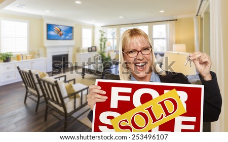 Happy Young Woman Holding Sold For Sale Real Estate Sign and Keys Inside Beautiful Custom Living Room.