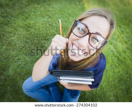 Fun Wide Angle Portrait of Pretty Young Woman with Books and Pencil Sitting in the Grass Outdoors.