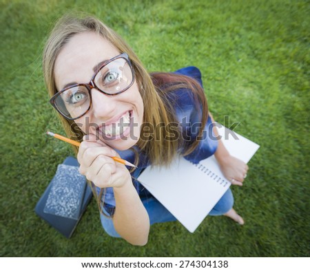 Fun Wide Angle Portrait of Pretty Young Woman with Books and Pencil Sitting in the Grass Outdoors.