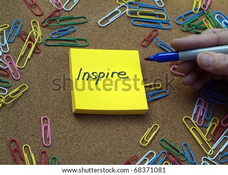 hand of person writing the word inspire on a yellow sticky note pad with bulletin board and colorful paperclips behind.