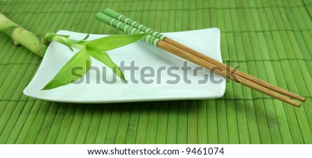 A green leafy bamboo shoot sits on a white plate with chopsticks on a green bamboo mat.