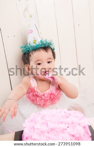 baby girl with icing on face looking down at her smashed pink roses birthday cake while sitting on floor white background with colorful hat