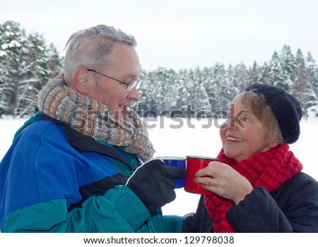 Elderly happy couple toasting with cups of warm drinks, outside in snow winter forest landscape