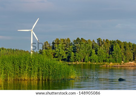 White wind turbine rotating in a sea shore green and blue landscape with evening sunlight and dark clouds - wind energy, sustainability and ecological concept background
