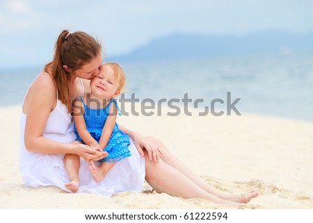 Young loving mother kissing her toddler daughter on white sand tropical beach