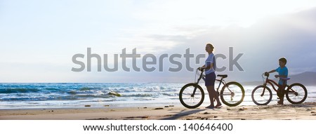 Panorama Of Mother And Son Biking On A Beach At Sunset