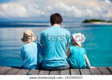 Back View Of Father And Kids Sitting On Wooden Dock Looking To Ocean