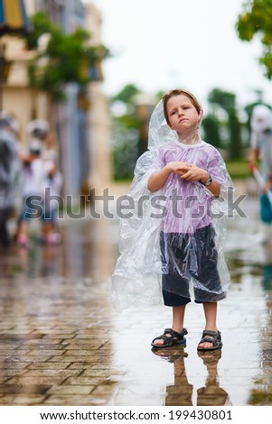Little boy in plastic transparent raincoat standing under the rain on summer day