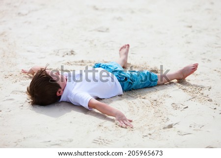 Little boy playing on tropical beach making an angel on white sand