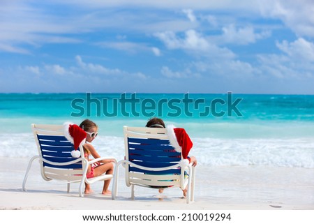 Two kids sitting on chairs with Santa hats at beautiful tropical beach enjoying Christmas vacation