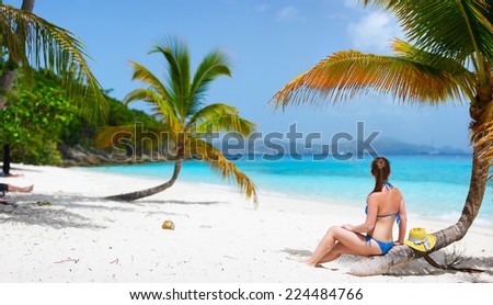 Back view of a young beautiful woman sitting on palm tree relaxing at white sand tropical beach