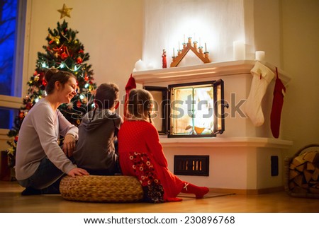 Family of mother and her two little kids sitting by a fireplace in their family home on Christmas eve