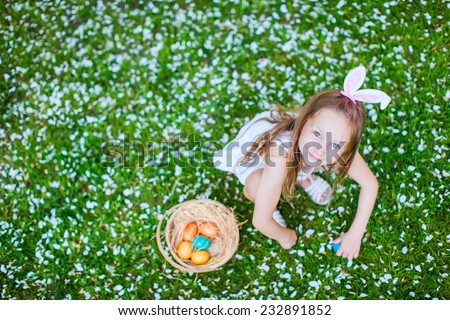 Top view of adorable little girl wearing bunny ears playing with Easter eggs on a grass covered with white flower petals on spring day