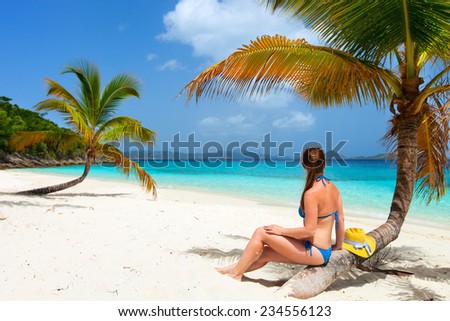 Back view of a young beautiful woman sitting on palm tree relaxing at white sand tropical beach