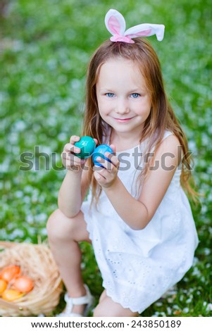Adorable little girl wearing bunny ears playing with Easter eggs on a grass covered with white flower petals on spring day