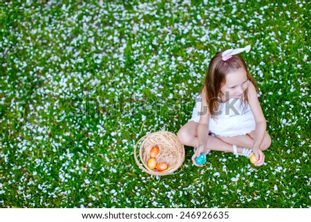 Above view of adorable little girl wearing bunny ears playing with Easter eggs on a grass covered with white flower petals on spring day