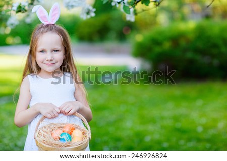 Adorable little girl wearing bunny ears holding a basket with Easter eggs in a blooming garden on spring day