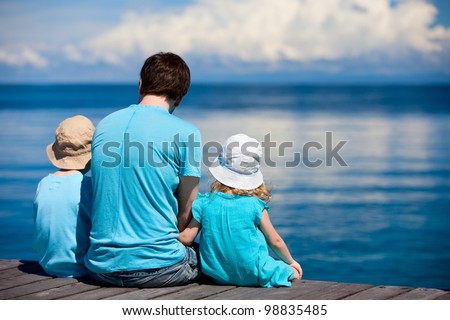 Back view of father and kids sitting on wooden dock looking to ocean