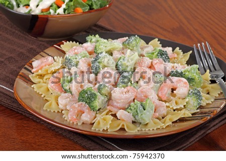 Shrimp, pasta and broccoli with a sauce on a plate with a salad in background