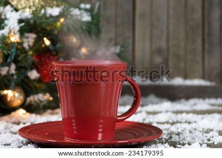 Red coffee cup with steam on snowy wooden deck with Christmas decorations in background
