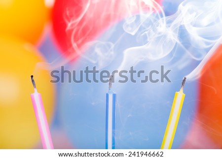 Three colorful birthday candles and smoke with balloons in background