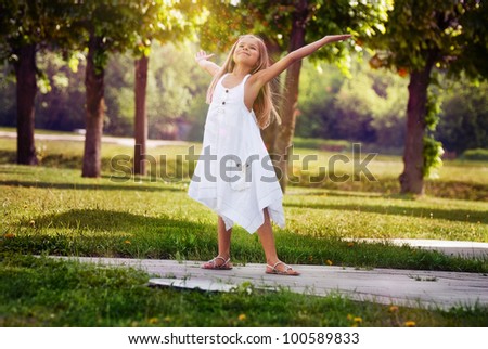 Cheerful and happy little girl with arms outstretched summer  in a white dress outdoors in a park smiles sweetly.