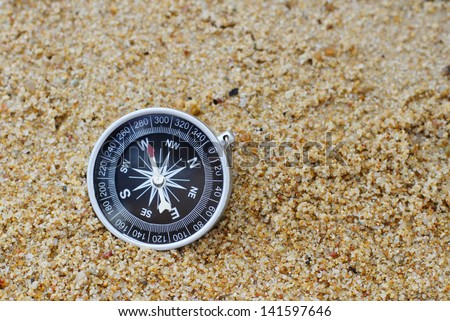 An old style compass pointing west resting on a sandy beach.