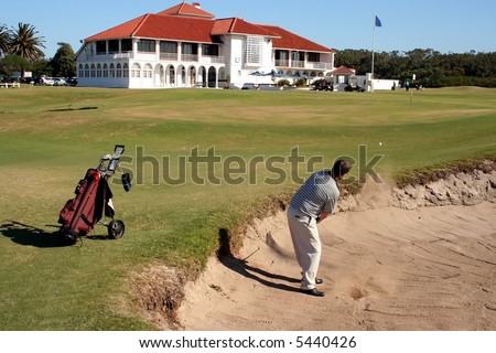 A golfer playing a stroke out of a sand trap with the club house in the background