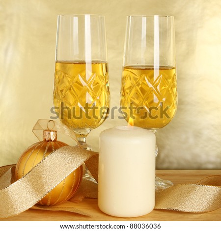 Two glasses of champagne, gold ribbon and Christmas bauble with a burning candle against a gold background