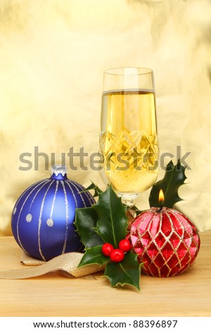 Christmas cheer, a glass of champagne with a blue bauble, decorative burning candle, gold ribbon and a sprig of holly against a gold background
