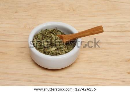 Dried tarragon herb leaves in a ramekin with a wood spoon on a wooden board