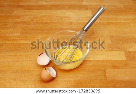 Eggs and a whisk in a glass mixing bowl with egg shells on a wooden kitchen worktop