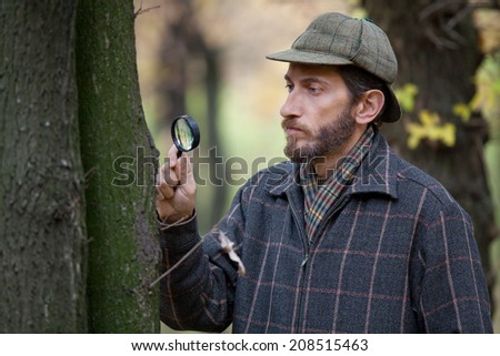 Man detective with beard wearing cap and plaid jacket considers through magnifying glass tree in autumn forest