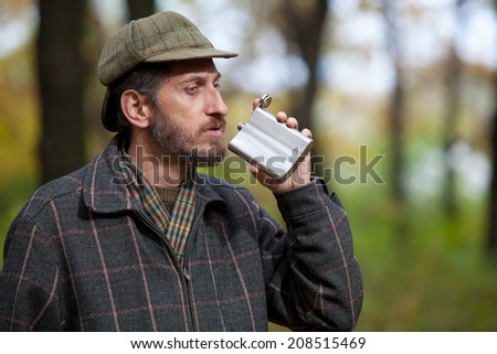 Man with a beard wearing cap and plaid jacket brings to mouth metal flask in autumn forest