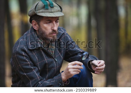 Imposing man detective with a beard wearing a cap and a plaid jacket considers through magnifying glass fragment of a tree in autumn forest