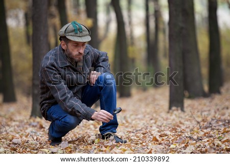 Man detective with a beard wearing a cap and plaid jacket considers through magnifying glass trail in autumn forest