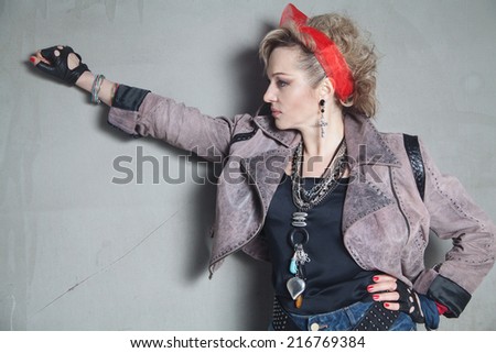 Beautiful blonde woman in rock style stands in profile near concrete wall with a raised hand